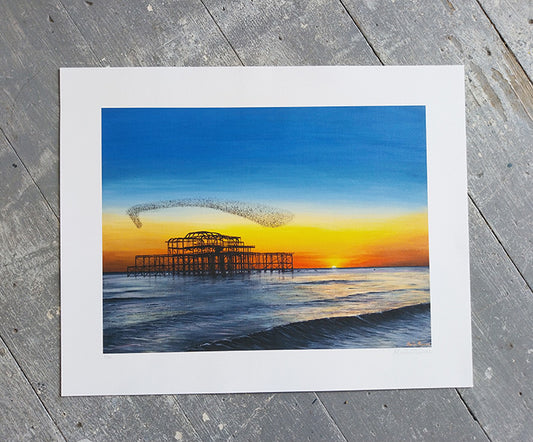 Art print of a sunset over Brighton west pier with a white border on a wooden surface.