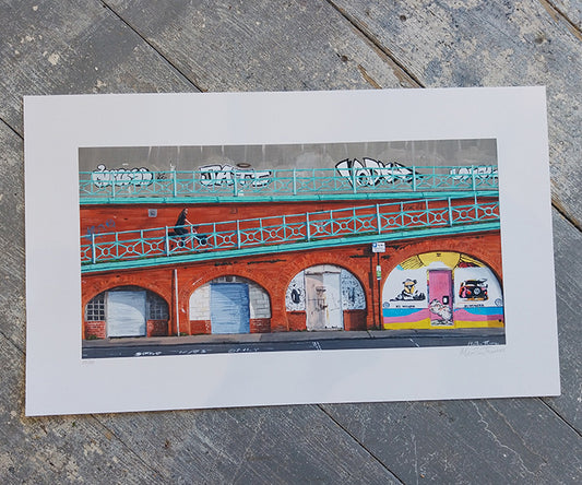 Art print of Arches in Brighton with Colorful mural on a brick wall with graffiti art, displayed on a wooden surface.